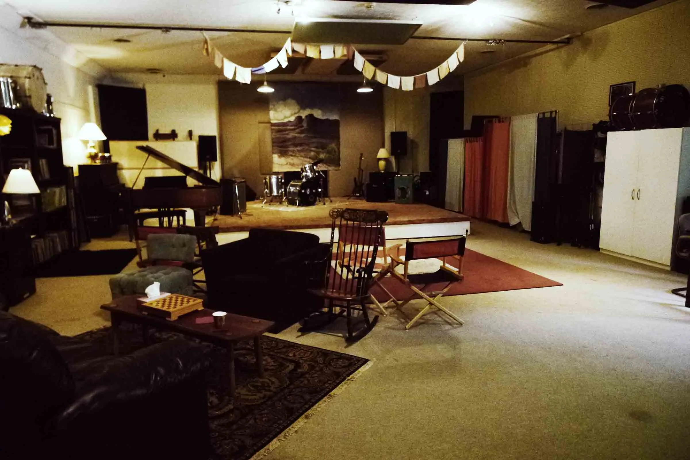 Cozy music studio with a drum set on a stage, grand piano, seating area with chairs and couches, and warm lighting.