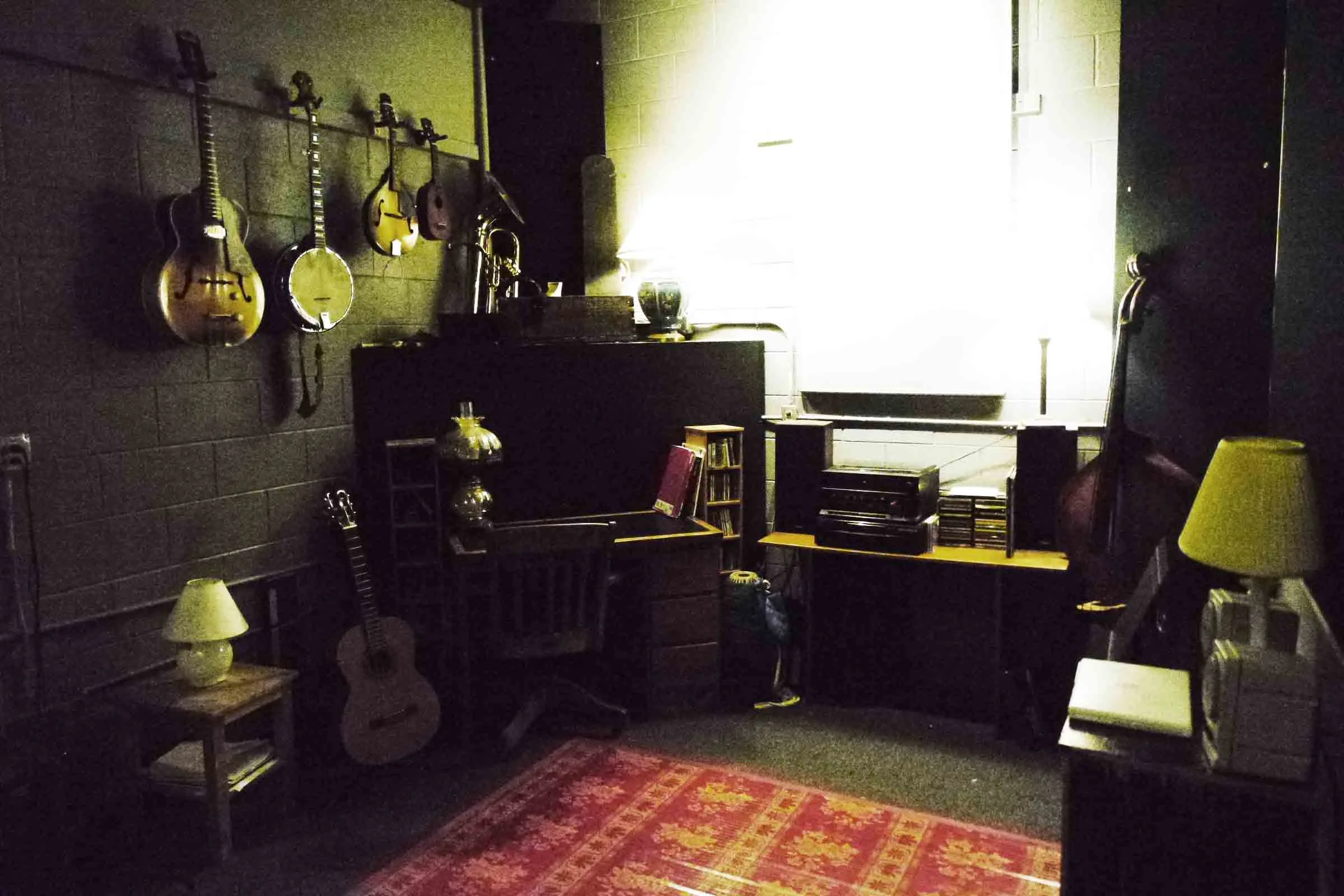 Dimly lit room with various musical instruments including guitars, banjo, mandolin, trumpet, and upright bass, along with a desk, lamps, and audio equipment.