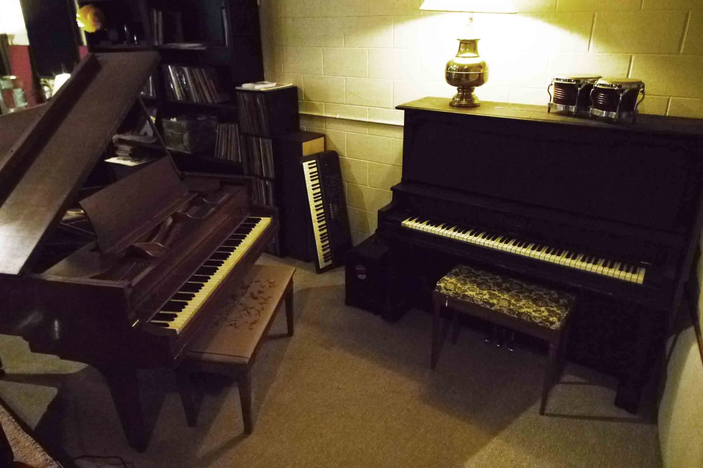 Cozy music room with a grand piano, an upright piano with a patterned bench, a keyboard leaning against a wall, and a lamp on top of the upright piano.